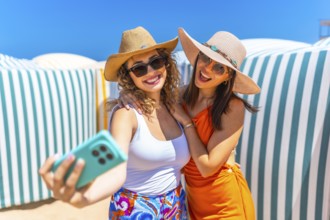 Two cheerful women wearing summer hats and sunglasses are taking a selfie on a sunny beach with