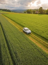 A car drives on a rural road through green fields, a village in the background, Deer E- CarSharing,