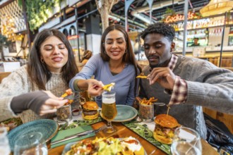 Three happy friends enjoying a meal together, sharing nachos, burgers, and fries at a restaurant