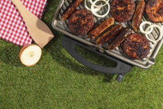Barbecue with sausages and burgers next to a red and white chequered cloth on a green lawn