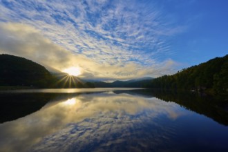 The early morning shows sunbeams through clouds reflecting on a calm lake in a mountainous