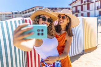 Two smiling women with sun hats and sunglasses taking a selfie on a sunny beach with colorful