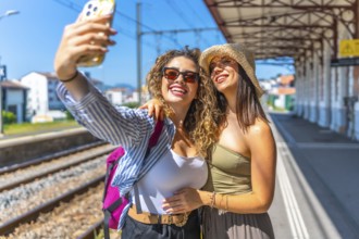 Two happy female tourists taking a selfie with smartphone at a train station, enjoying their summer