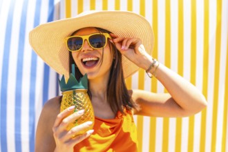 Young woman with sunglasses and a straw hat, enjoying a tropical pineapple juice on a colorful