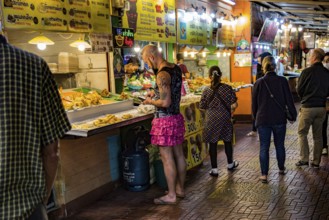 Tatooed man in pink skirt and flip flops ordering food from a street vendor in the Night Market in