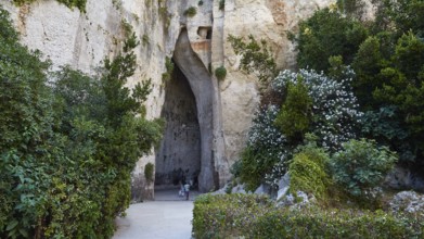 Cave Ear of Dionysus, Overgrown cave entrance with lush vegetation and rocks, people in the