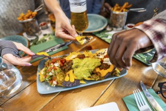 Hands grabbing nachos from plate with guacamole, cheese, jalapenos, and pico de gallo, served on