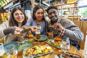 Three cheerful multi ethnic friends are enjoying a meal together, sharing nachos, burgers, fries,