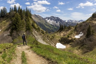 A hiker on a narrow path, surrounded by mountains and scattered snow under a blue sky in