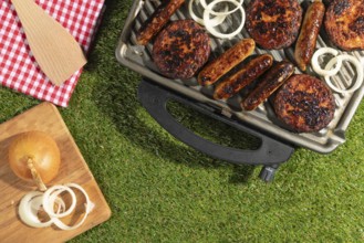 Sausages and burgers on a grill with onions on a wooden board on a green lawn
