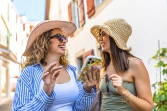 Two happy women wearing summer hats and sunglasses are using a smartphone and laughing while