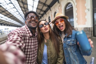 Three happy young tourists are taking a selfie on a train station platform, excited for their next