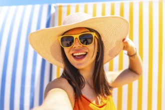 Young woman with sunhat and sunglasses taking a selfie on a striped background at the beach during