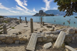 Ancient ruins in front of a rocky coast with a view of the clear sea and a small, mountainous