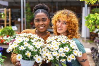Two happy multi ethnic female garden center workers holding pots of white daisies smiling at the