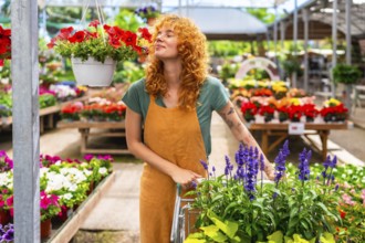 Redhead woman gardener smelling red petunias while pushing a cart in a colorful greenhouse full of