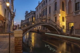 Small pedestrian bridge arching over canal in venice, italy at twilight with light trails