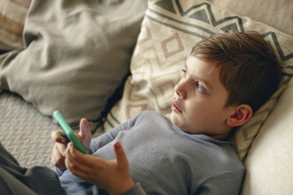 A boy holds a smartphone in his hands, relaxing on the couch, in a cozy living room, natural light,