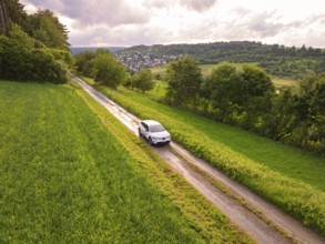 Car on narrow path between green meadows, view of village in the background, Deer E- CarSharing,