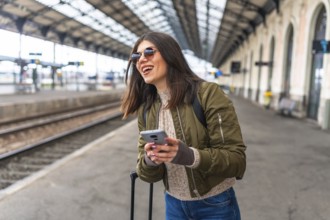 Young woman tourist using her smartphone while waiting for a train at the railway station, smiling