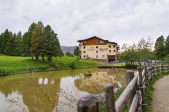 Building at a pond with surrounding trees and a wooden fence under a slightly cloudy sky, Alpe di