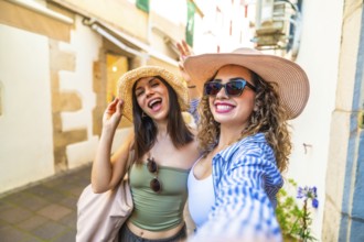 Two joyful women with straw hats and sunglasses are taking a selfie with a cheerful expression,