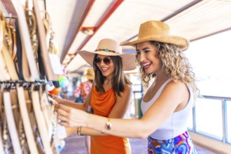 Two smiling women wearing summer hats and sunglasses are choosing souvenirs at a seaside shop while