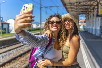Two female tourists with backpack and straw hat are taking a selfie with smartphone at a train