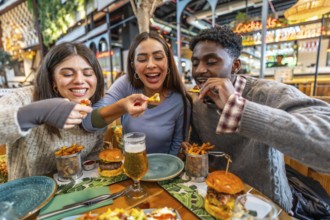 Three cheerful multi ethnic friends sharing nachos and burgers, enjoying a meal together at a