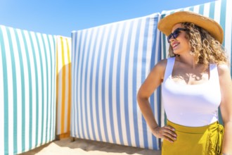 Young woman with curly hair, sunglasses and straw hat posing on beach with colorful striped cabanas