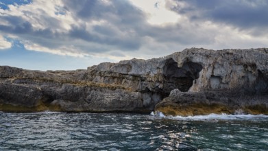Dramatic rocky coastline with natural arch and cloudy sky over the sea, boat trip around the island