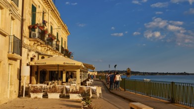 Cosy restaurant on a promenade with sea view. People walking in sunny weather, Ortigia Island,