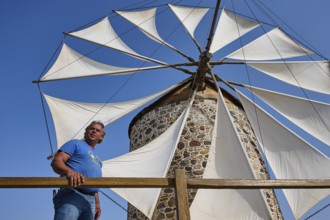 A man stands in front of a windmill with white sails under a clear blue sky, Windmill, Antimacheia,