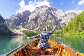 A man sits in a wooden boat and spreads out his arms as he heads towards an impressive mountain,