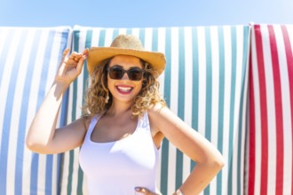 Cheerful young woman with curly hair, wearing sunglasses and a straw hat, smiles while posing