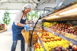 Smiling senior saleswoman wearing gloves, arranging vibrant fresh fruits in a supermarket display