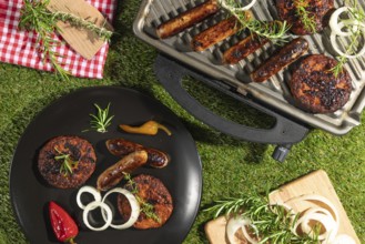 Grilled sausages and burgers on a barbecue and plates with rosemary on a green lawn
