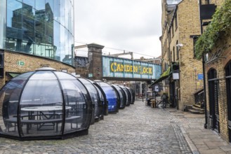 Transparent dining pods line a cobblestone street beneath the camden lock bridge in london,