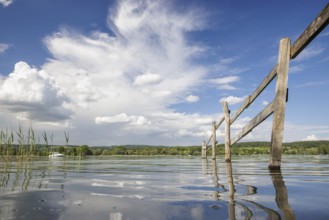 A wooden fence stretches into the calm waters of a lake, surrounded by a wide sky and white clouds,
