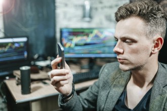 Man focused on a smartphone in an office with trading charts on monitors