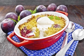 Crumble with plum and ice cream in a red brazier with spoons on a napkin, plums and mint on dark