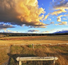 The huge cloud lit yellow-orange rays of sunset. Cozy wooden bench by the side of the road in