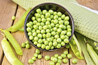 Green peas in a brown bowl, napkin, pea pods on the background of wooden boards
