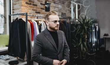 Man wearing sunglasses and a coat stands inside a chic clothing store with trendy decor