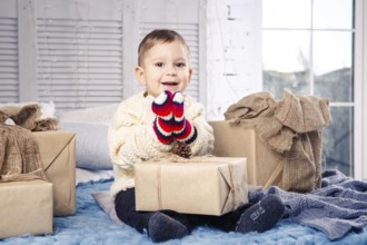 Little funny playful boy a child sits on a bed on Christmas day with gift boxes in white wool