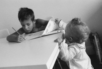 A boy sitting at the table and doing his homework. His curious baby sister is standing nearby and