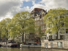 Building fronts along a canal with boats in the water, surrounded by green trees and a cloudy sky,