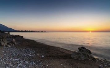 A peaceful sunset over the ocean with silhouette of mountains and pebble beach iwth rock boulders