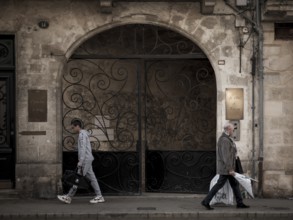 Individuals walk past an arched entranceway with ornate iron gates in central Bordeaux, France,