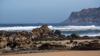 Rocky coast with large stones, behind it the sea with gentle waves under a clear blue sky, Praia de